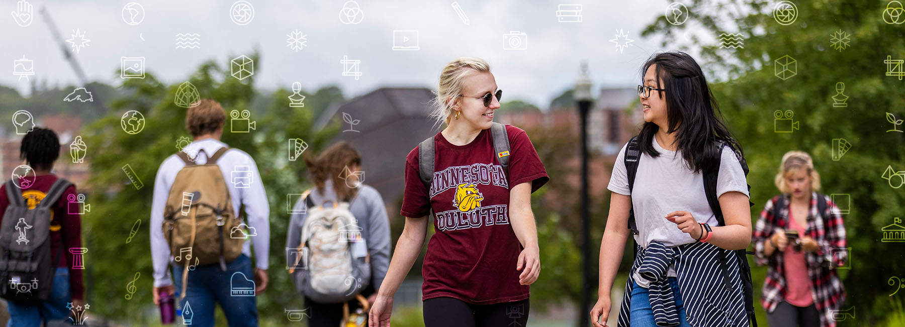 two female students walk and talk side by side outside on campus with other students and Weber Music Hall in the backdrop. Artistic overlay of icons that depict the areas of study within the College of Arts, Humanities, and Social Sciences.