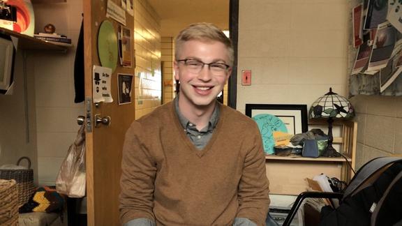 Smiling blonde man wearing glasses and sweater in an office setting. 